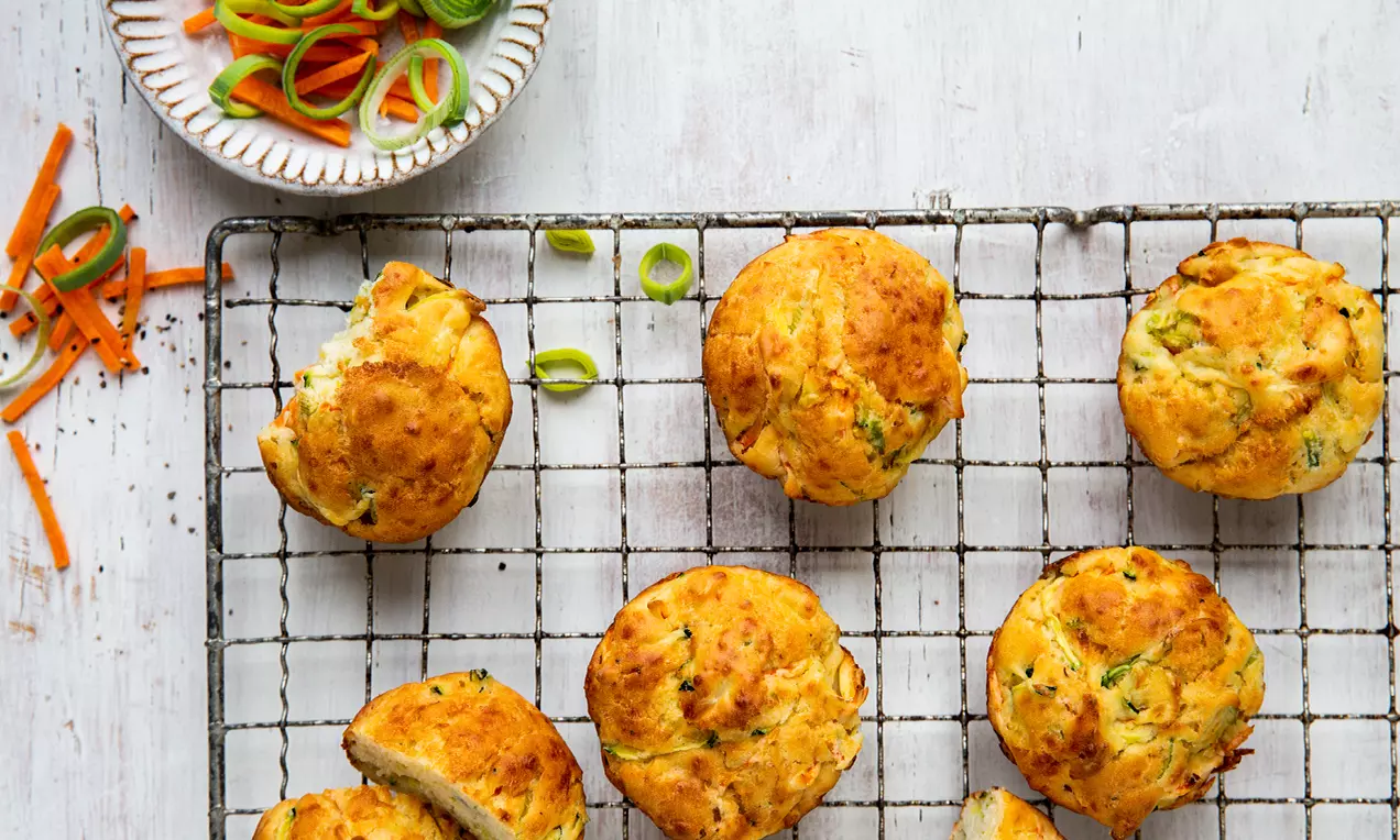 Goldbraune Gemüsemuffins mit leichter Kruste auf einem schwarzen Gitterrost, daneben bunte Lauch- und Karottenstreifen in einer weißen Schale