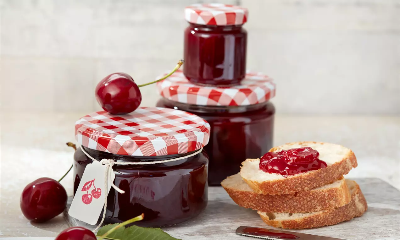 Confettura di ciliegie rosso intenso in barattoli di vetro con coperchi a quadretti bianchi e rossi, accanto a fette di pane dorato e ciliegie fresche