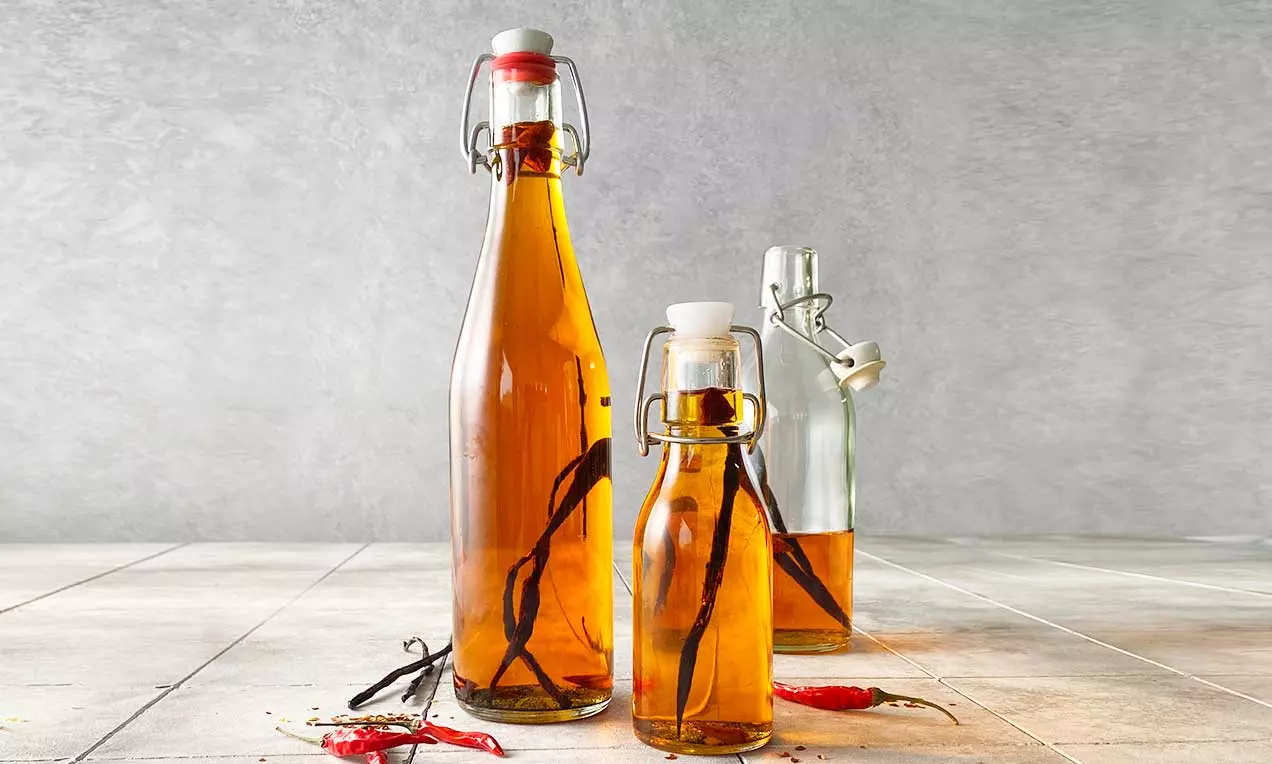 Bouteilles transparentes remplies d'huile ambrée avec des gousses de vanille et des piments rouges sur un fond clair