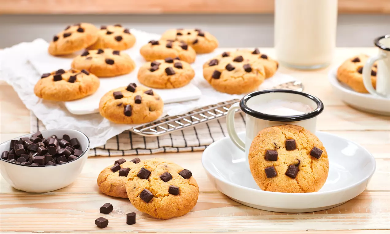 Biscotti dorati e rotondi con gocce di cioccolato scuro, disposti su una griglia e un piatto accanto a una tazza di latte e ciotole con cioccolato