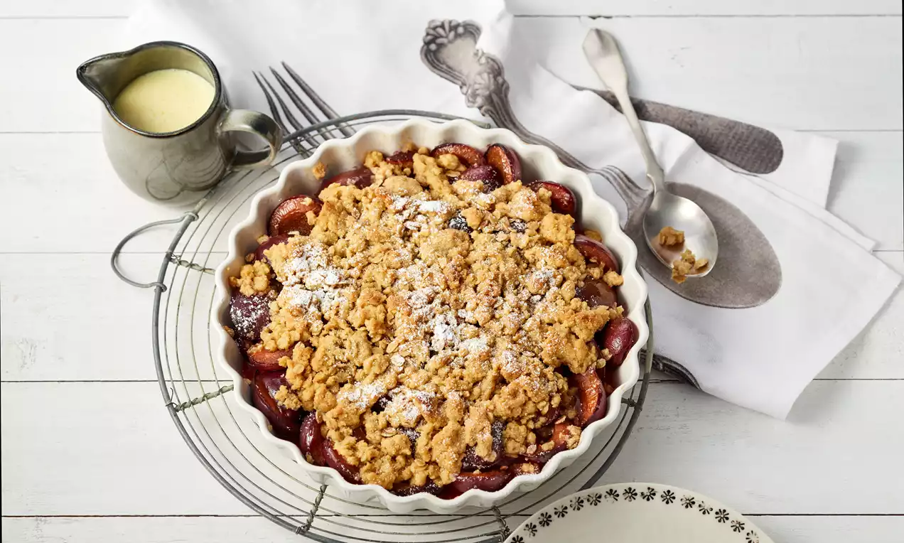 Zwetschgen-Crumble mit goldbrauner Kruste und Puderzucker in einer weißen Form auf einem Metallgitter, daneben Kanne mit Vanillesauce