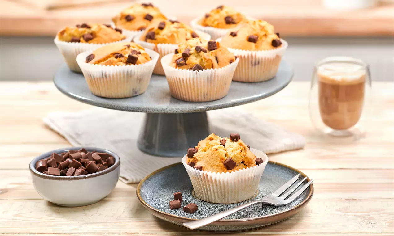 Muffin dorati e soffici con gocce di cioccolato su alzata grigia, un piatto con forchetta e cioccolato a pezzi accanto a un caffè in bicchiere